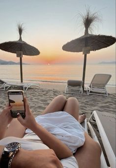 a man laying on top of a beach next to the ocean holding a cell phone