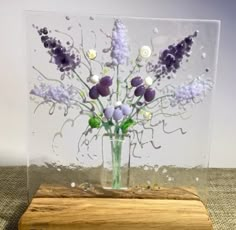 a glass vase filled with purple flowers on top of a wooden block in front of a white wall