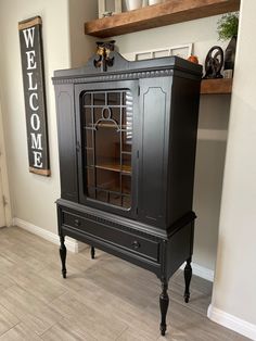 a black cabinet sitting on top of a tile floor next to a wooden shelf with a welcome sign above it