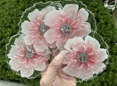 a person holding three pink flowers in front of a green plant with glitter on the petals