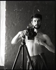 a shirtless man holding a camera in front of a tripod while standing next to a wall