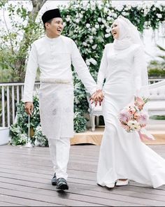 two people dressed in white holding hands and walking on a deck with flowers behind them