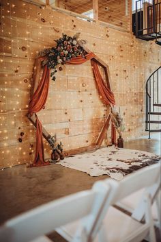 a wedding ceremony setup with orange draping and flowers on the wall, surrounded by white chairs