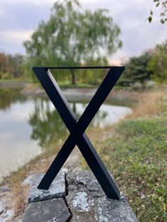 a metal x sign sitting on top of a stone wall next to a lake and trees