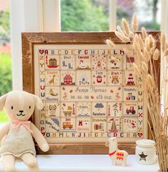 a stuffed animal sitting next to a framed cross stitch pattern on a window sill