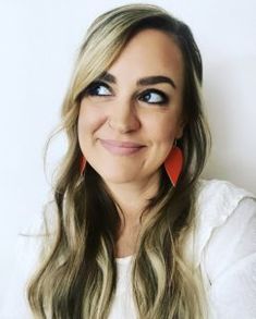 a woman with long blonde hair and blue eyes smiles at the camera while wearing red earrings