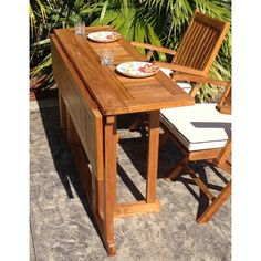 a wooden table with two chairs and plates on it, in front of some plants