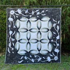 a black and white quilt hanging on a fence in front of some bushes with flowers