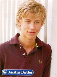 a young man with blonde hair wearing a maroon shirt and smiling at the camera while leaning against a white wall