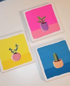 three square paintings with small potted plants on them in different colors and shapes, one is pink, the other is blue