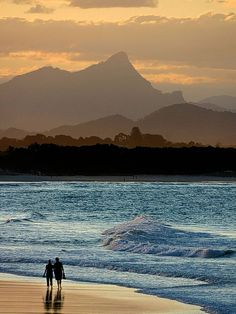 two people are walking on the beach at sunset with mountains in the backgroud