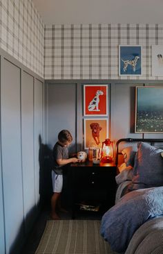 a young boy standing next to a night stand with a lamp on it in a bedroom