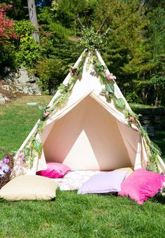 a teepee tent with pillows and flowers on the ground in front of it, surrounded by greenery