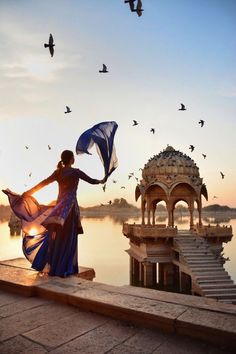 a woman holding an umbrella standing next to a body of water with birds flying around her