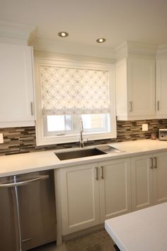 a kitchen with white cabinets and stainless steel dishwasher next to a window that has roman shades on it