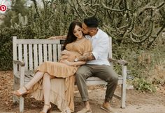 a man and woman sitting on a bench in front of some trees with their arms around each other