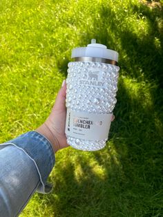 a person holding up a white and silver coffee cup in their hand on the grass