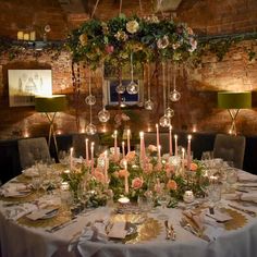 a table set with candles and flowers in front of a brick wall, surrounded by greenery