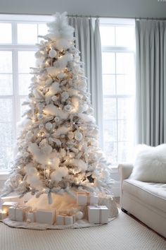 a white christmas tree with presents under it in front of a large window and curtains