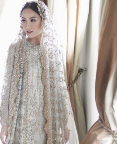 a woman in a wedding dress and veil standing next to a window with curtains behind her