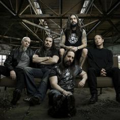 five men are sitting on a couch in an industrial building, one is wearing a black shirt