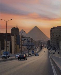 cars are driving down the road in front of some buildings and pyramids at sunset