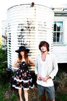 two people standing next to each other in front of an old metal silo and building