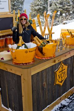 a woman standing behind a wooden counter filled with bottles and buckets on top of snow covered ground