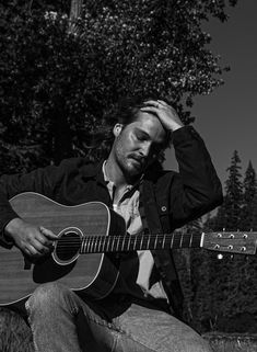 a man sitting in the grass holding a guitar and looking at his head with one hand