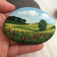 a hand holding a painted rock in front of a field with red flowers and trees