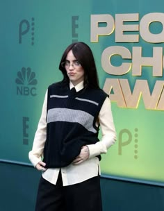 a woman with glasses standing in front of a green wall wearing a black and white sweater