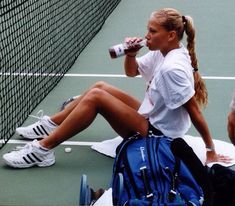 a woman sitting on the tennis court drinking from a water bottle while holding a backpack