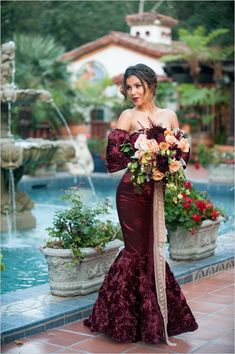 a woman in a long dress standing next to a fountain with flowers on her shoulder