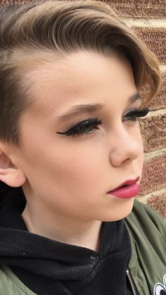 a woman with short hair wearing black eyeliners and a green jacket standing in front of a brick wall