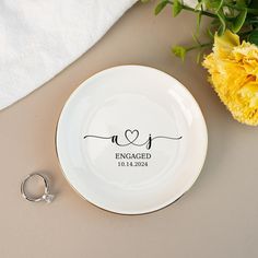 an engagement ring sits next to a plate with the word engaged on it and a yellow carnation