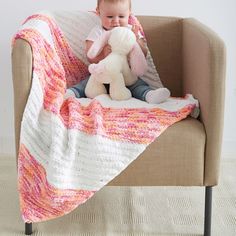 a baby sitting on a couch holding a teddy bear under a pink and white blanket