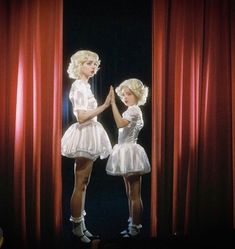 two women in white dresses standing on stage with red curtains behind them and one holding her hand up to the other side