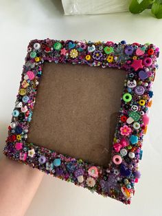 a hand holding a small frame made out of buttons and other things on the table