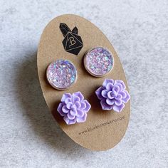 two purple flower shaped earrings sitting on top of a piece of brown paper with glitter