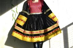 This is a richly textured traditional Afghan dress and Its main fabric is velvet, in a deep, luscious black that gives it a luxurious sheen. The dress features vibrant patterns and colors on the chest which is hand knotted and cuffs, with a geometric design that includes shades of red, orange, and green. A standout feature is the elaborate gold embroidery and trim that adorn the hem and cuffs, adding a regal touch. Fitted Black Dress For Festivals, Black Dress With Dabka In Traditional Drape, Traditional Black Festival Dress, Black Traditional Drape Dress For Festivals, Traditional Black Dress For Festival, Black Bohemian Dress With Traditional Patterns, Black Dresses With Traditional Drape For Transitional Season, Black Festive Dress For Festivals, Black Dress For Festive Transitional Season