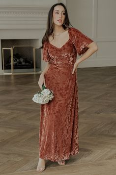 a woman standing in a room wearing a brown dress and holding a white bridal bouquet