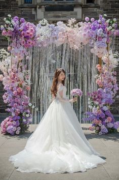 a woman in a wedding dress standing under a floral arch with purple and white flowers