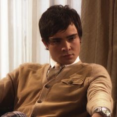 a young man sitting on top of a couch in front of a window wearing a sweater and tie