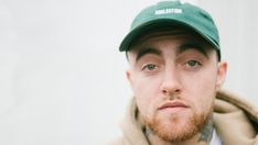 a man with a beard wearing a green hat and coat looking at the camera while standing in front of a white wall