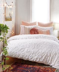 a bed with white comforter and pillows on top of it next to a potted plant