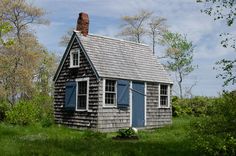 a small gray house sitting on top of a lush green field next to tall trees