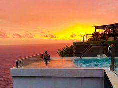 a man is sitting on the edge of a swimming pool at sunset, overlooking the ocean