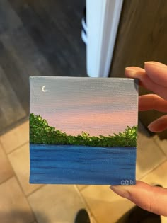 a person holding up a small painting in their hand with the sky and trees painted on it