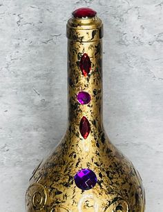 a gold colored vase with red and purple stones on the top, sitting against a white wall