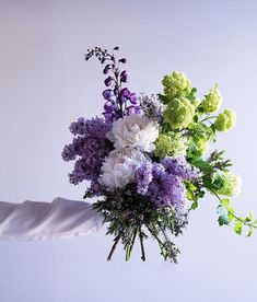 a bouquet of purple and white flowers is held up by someone's hand in front of a white wall